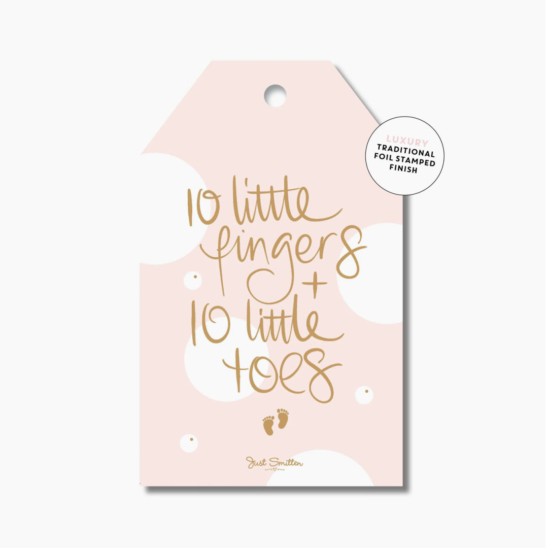 A pink gift tag with gold foil-stamped text reading '10 little fingers + 10 little toes' and a small pair of footprints below the text.