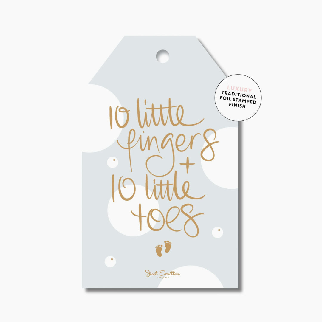 A blue gift tag with gold foil-stamped text that reads &#39;10 little fingers + 10 little toes&#39; and the &#39;Just Smitten&#39; logo at the bottom.