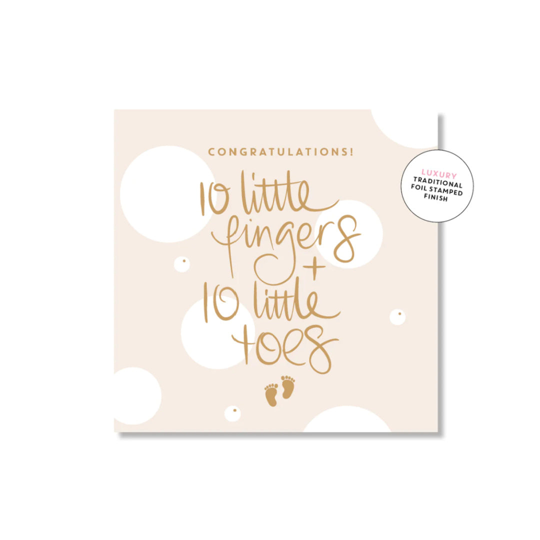 A greeting card with 'CONGRATULATIONS! 10 LITTLE FINGERS + 10 LITTLE TOES' written in gold foil on a neutral background with gold dots.