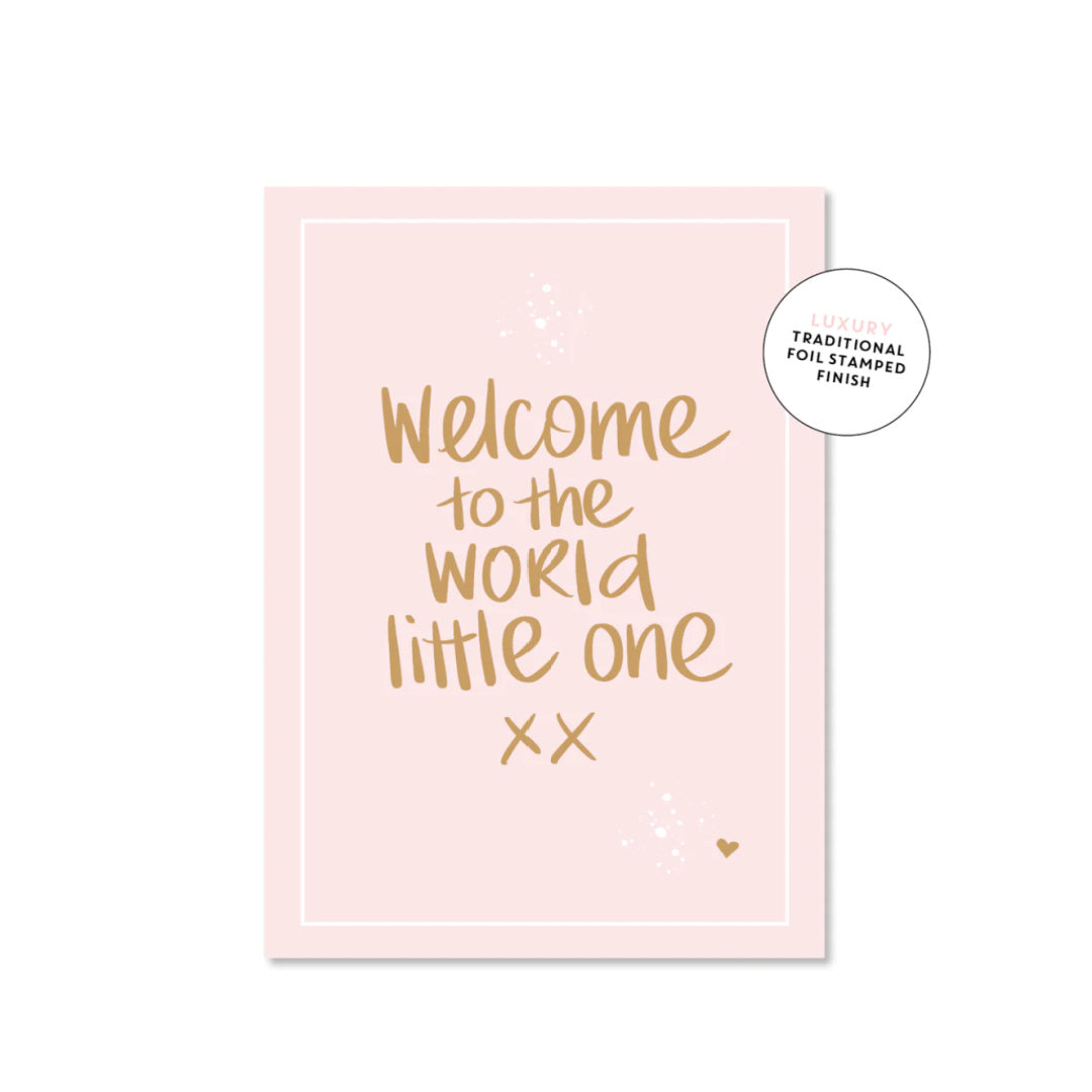 A luxury greeting card with 'Welcome to the world little one' printed in gold foil on a pink background, with a heart symbol at the bottom right corner.